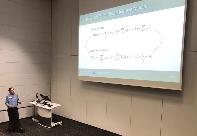 Photograph of Georgia standing at the front of a lecture theatre featuring towards a slide on a big projector with the title ‘local control for the Bose Hubbard model’ and equations labelled ‘where it started’ and ‘where we are going’.