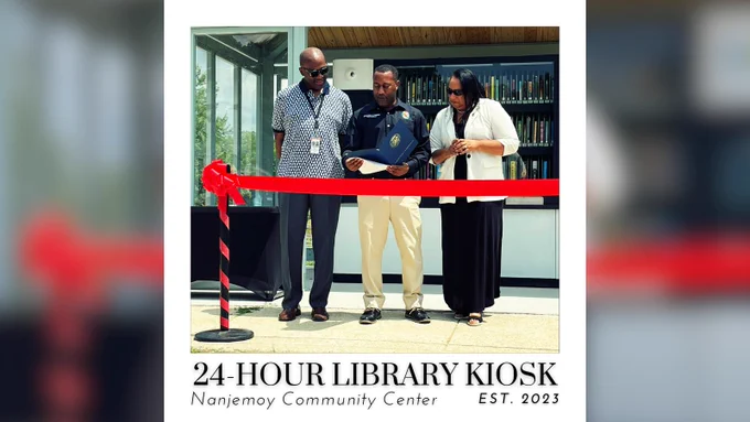 Charles County Public Library's Executive Director, KennethWayne Thompson, Commissioner Vice President Ralph Patterson, & Commissioner Thomasina Coates stand behind a red ribbon, as CVP Patterson reads a Citation at the 24-Hour Library Kiosk Grand Opening and Ribbon Cutting Ceremony. It is a bright, sunshiny day as they stand in front of the kiosk.