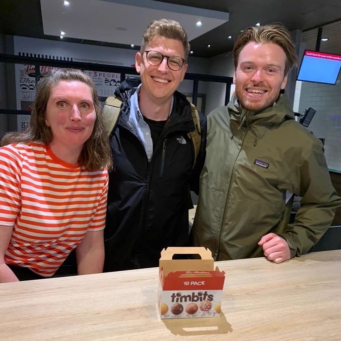 Three people smile with a box of timbits on the table.