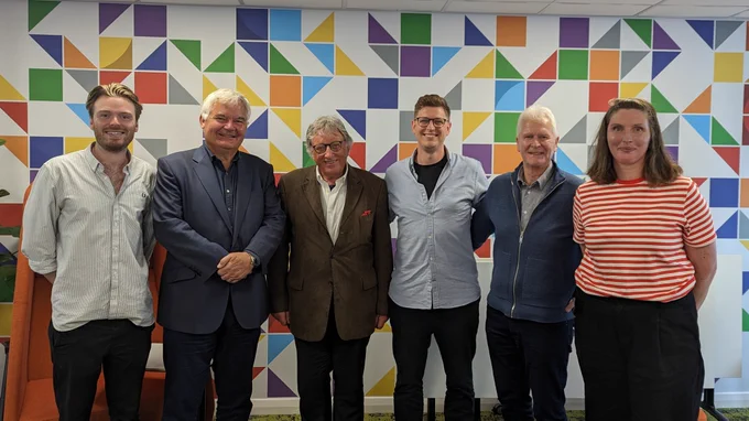 5 men and one woman stand in front of a colourful pattern wall smiling. 