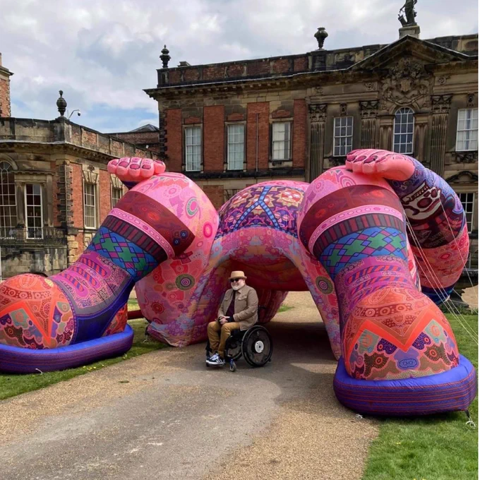 A man in a wheelchair is wearing a stone coloured jacket, trilby hat, sunglasses. He is in front of a large colourful sculpture that he created of what is meant to be a person laid down with their legs open. The picture has been taken outside in front of a stately home type building. 