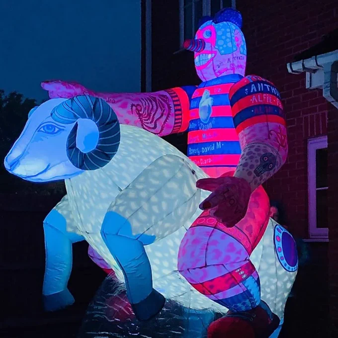 A picture taken at night time of a colourful inflatable sculpture that is lit up inside. The abstract sculpture is of a person with tattoo like drawings on their body, wearing red and blue striped top and shorts and riding on the back of a ram.