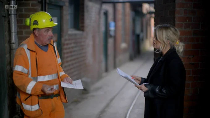 Pete in a full suit of high-vis orange and hard hat, talking with Emily outside, holding documents