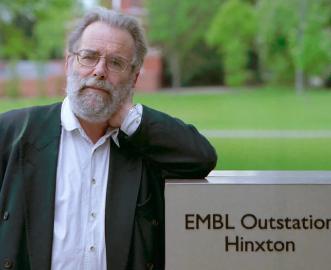 man leaning against a sign reading "EMBL Outstation Hinxton"