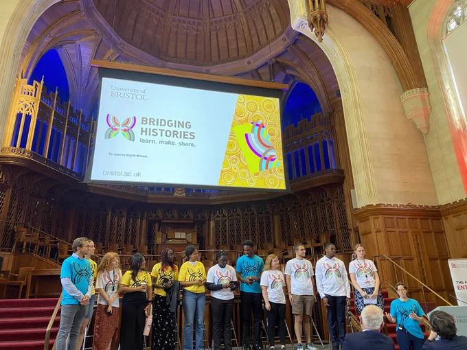 Dr Joanna Burch-Brown and Summer School students on stage in the Great Hall, Wills Memorial Building talk about Bridging Histories before a projector displaying the project logo.