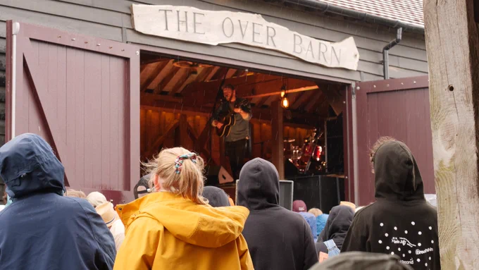 Plested playing the guitar and singing in the wooden barn, A sign above the door says "the over barn" engraved in a wooden plank. It is raining and the audience can be seen wearing raincoats