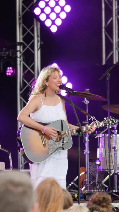 Cate on stage playing a semi-acoustic guitar, wearing all white. The stage is illuminated in purple.