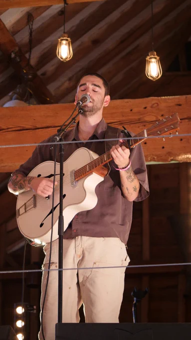 Adam Melchor playing guitar in the wooden barn wearing a brown shirt and sand coloured trousers. Above him, two glowing pendant lights can be seen.
