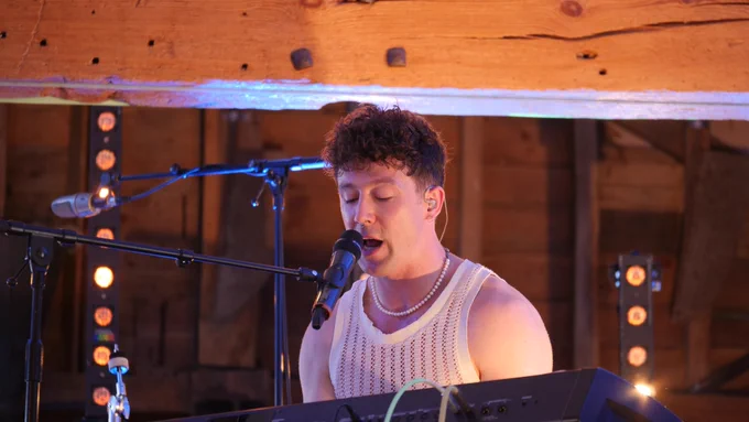 Moncrieff playing the piano in the wooden barn stage. A wooden beam can be seen above him. He's wearing a white top.