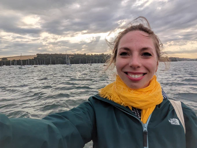 Selfie of me on a boat with the ocean and the shore in the background, evening light filtering through the clouds and playing on the waves. It's impossibly picturesque 