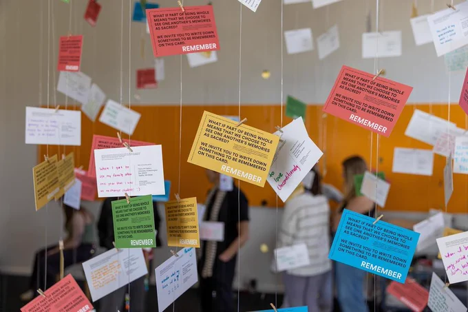 several people are standing in a room with colorful notes hanging from the ceiling, part of an exhibition by artist Daniel Alexander Jones