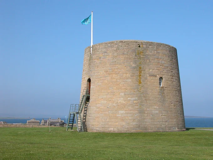 Hackness Martello Tower. The image shows a circular stone structure in the right middle of the frame. It is accessed by external metal stairs on its left and surmounted by a flagpole with a light blue flag. The foreground is grassy and there are some buildings and the sea in the background. The sky is blue.