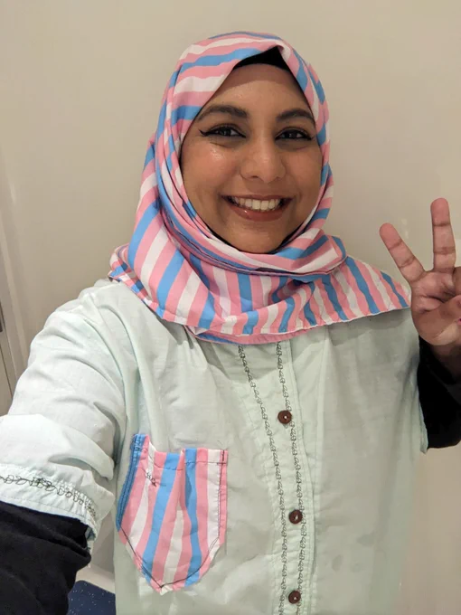 Photo of Hafsa showing a peace sign, wearing a trans flag hijab and a shirt with a pocket that has the trans flag on it