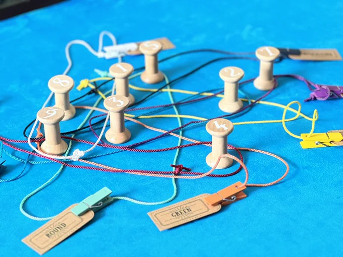 Spools labelled 1-8 are placed on a blue surface. Coloured looped threads are placed around the spools such that they cross over one another. At the end of each loop is a peg holding a word and a card ticket. The words ‘round’ and ‘green’ are in focus