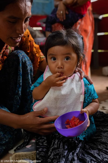 A child is looking at the camera while she is eating a fruit from a bowl with her fingers while a woman holds her.