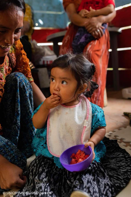 A child is looking at a woman while she is eating a fruit from a bowl.
