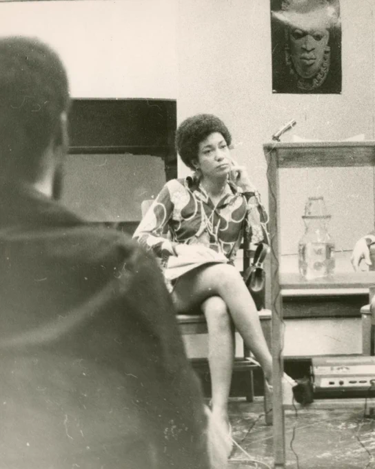 A black and white sepia tone photograph of poet June Jordan. Jordan sits in a chair with her proper left arm resting against her cheek. An unidentified person sits with their back towards the camera.