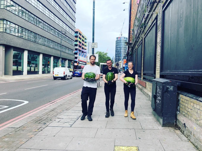 Three people walk down a London street, each carrying a large watermelon.