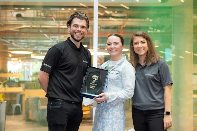 A photo of three adults smiling with the middle adult holding a trophy. There is a colourful glass back drop behind them. 