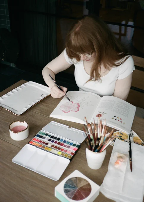 Sitting at an outdoor patio table on a sunny day, BDH paints a pink and red rose in Sarah Simon’s new watercolor workbook, “Flowers, Feathers, & Animal Friends.” She wears a white t-shirt, glasses, and her hair down.