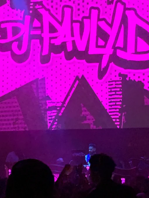 Pauly D inside a club in front of a crowd, a giant screen behind him with “DJ Pauly D” written in purple.