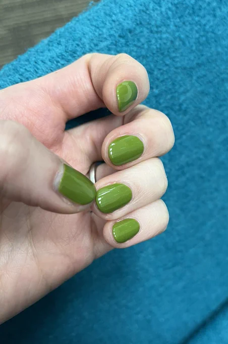 Manicure shot in front of teal upholstery. Polish is a dark avocado shade, approaching olive, but vibrant.