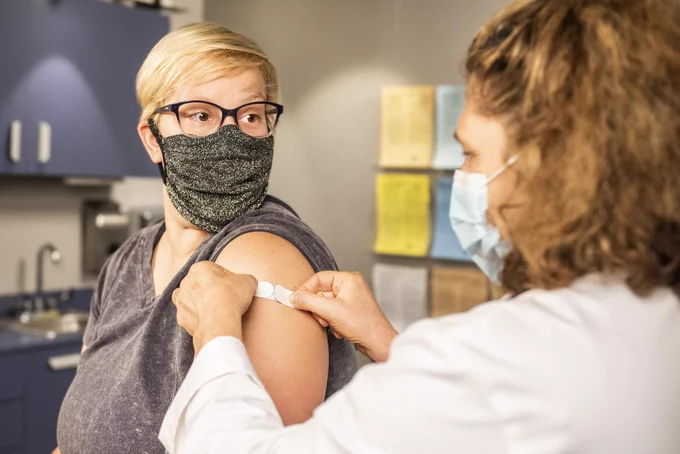 Woman wearing face mask has plaster applied by a clinician following Covid-19 vaccination.