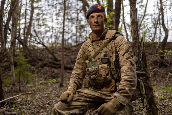 A white British Army officer is sitting in the woods with the background being slightly out of focus. He is wearing a green army uniform and a beret.