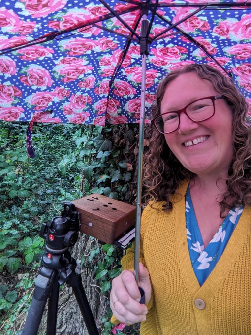 Woman in yellow cardi stands under a pink and yellow umbrella. There is a camera stand and wooden box camera beside her, and the green and brown of the forest behind that.
