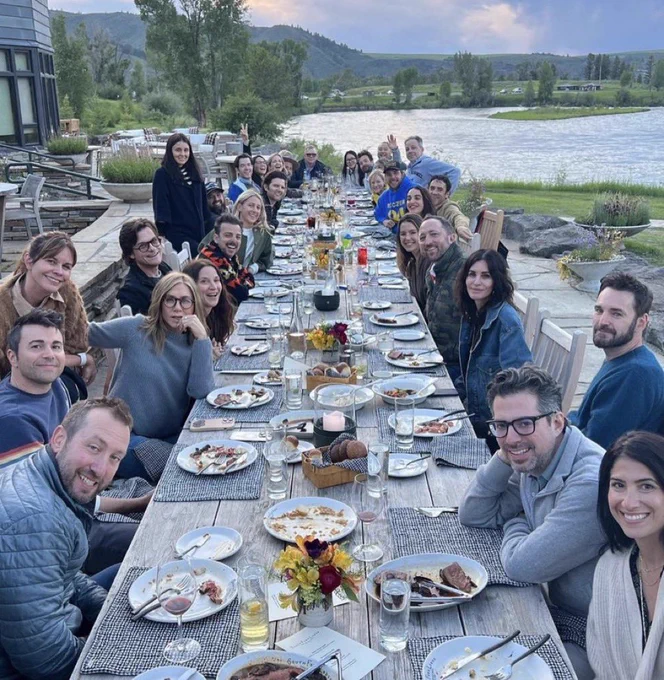 a bunch of (white) celebrities sitting at a long dining table near a lake, posing for a picture.