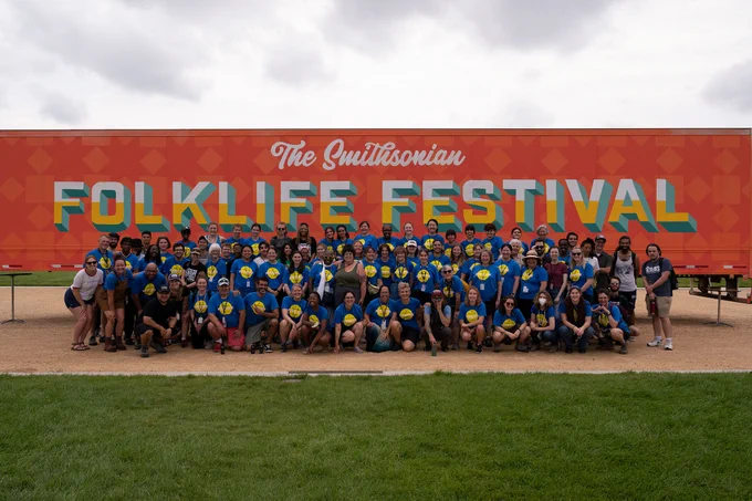 A large group of people in blue T-shirts stand in front of a large trailer, smiling at the camera. The trailer is painted with an orange background and light orange diamonds and squares. The text on the trailer reads “The Smithsonian Folklife Festival.”