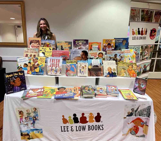 Lee & Low's Head of Educational Sales, Verónica Labarca, stands and smiles behind a table at the California Association of Black School Educators. The table holds a plethora of Lee & Low titles.