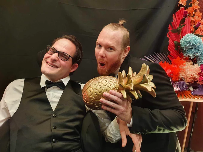 Two men in formalwear pulling faces and holding a golden pineapple statuette