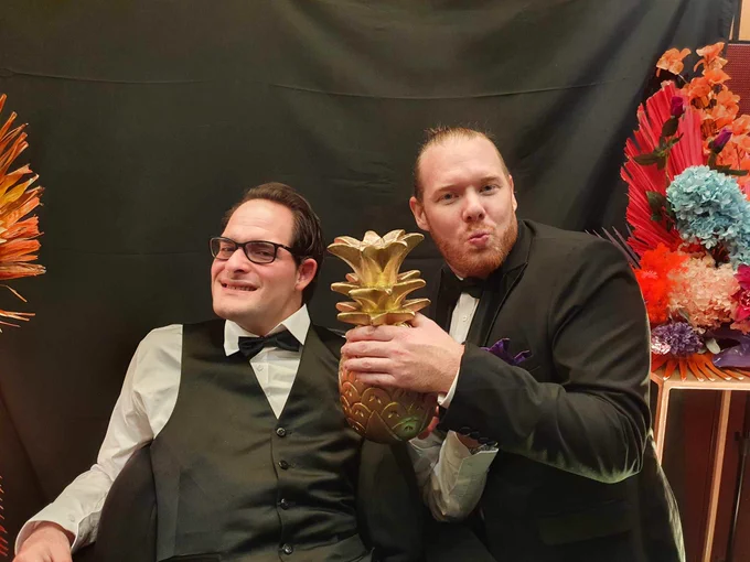 Two men in formalwear pulling faces and holding a golden pineapple statuette