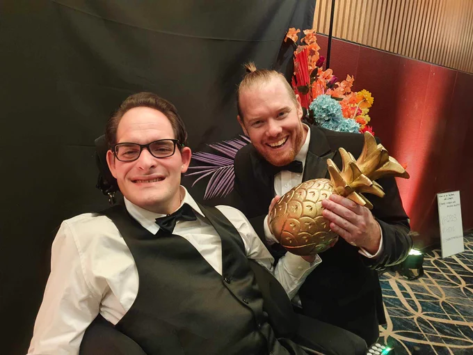 Two men in formalwear pulling faces and holding a golden pineapple statuette