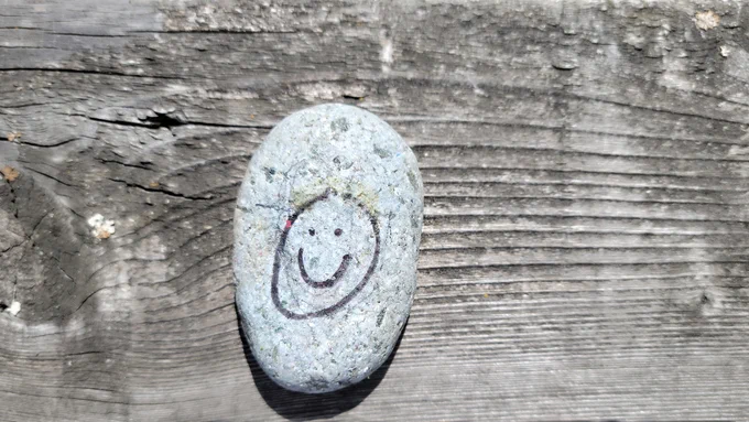 An ocean-polished stone sitting on top of a cedar fence. On the face of stone, a happy face is drawn in black marker. The reverse side of the stone has the artist's name (Niklas A.) and the word happy