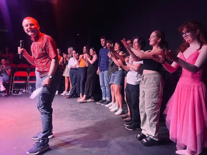 Early Dawn New Writing Night, Josh stands on the left of the image in a red checked shirt. Behind him are the writers and performers of Early Dawn