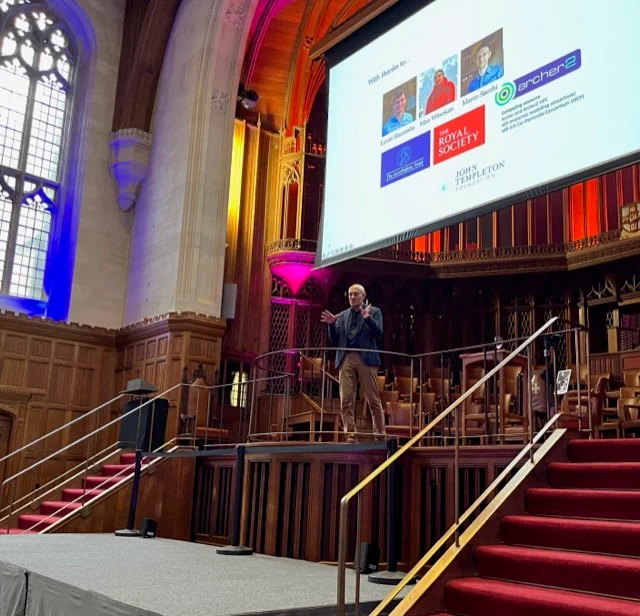 Professor Jim Al-Khalili standing speaking on stage in the Great Hall of the Wills Building at the University of Bristol. A Powerpoint presentation is showing on a large screen above Professor Al-Khalili.
