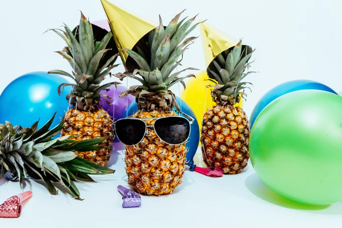 3 pineapples wearing party hats, one with sunglasses, and surrounded by balloons and party blowers.