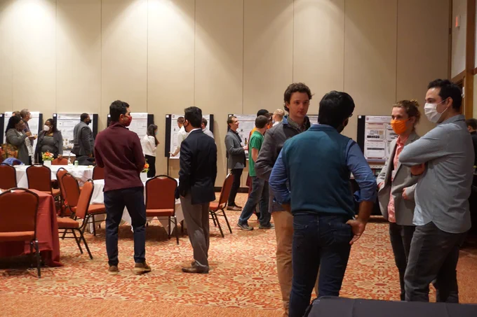 A group of people stand in a banquet hall, discussing poster displays. 