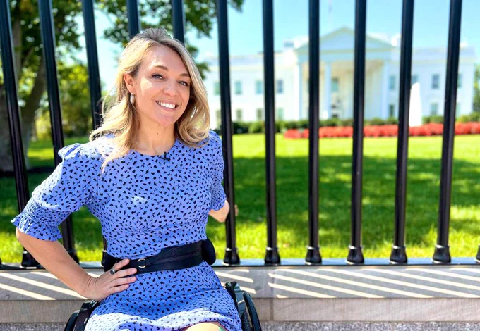 Sophie is outside the White House, in her manual wheelchair, smiling with long blonde hair, wearing a purple and black dotted dress.