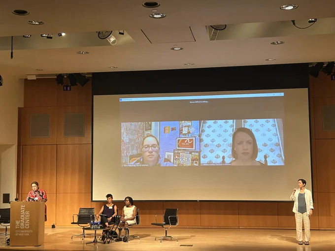 On the left, one woman on the podium amid spreads, two women sitting by her on stage, two women are projected on the screen behind them. ASL interpreter is standing on the right of the stage.