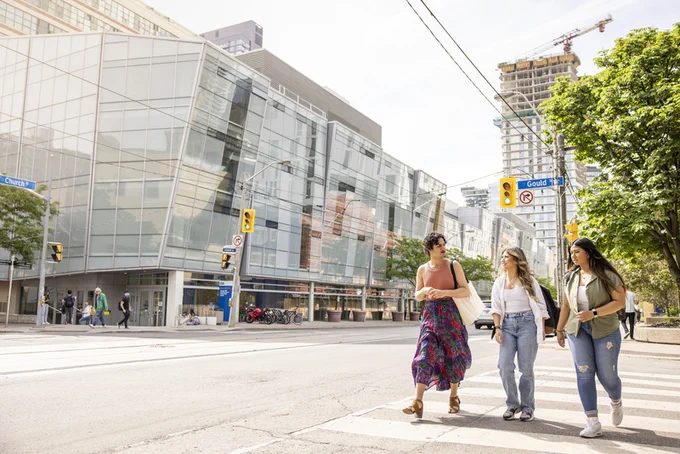 Three smiling TMU students chat as they use the crosswalk in front of the George Vari Engineering and Computing Centre on Gould Street.