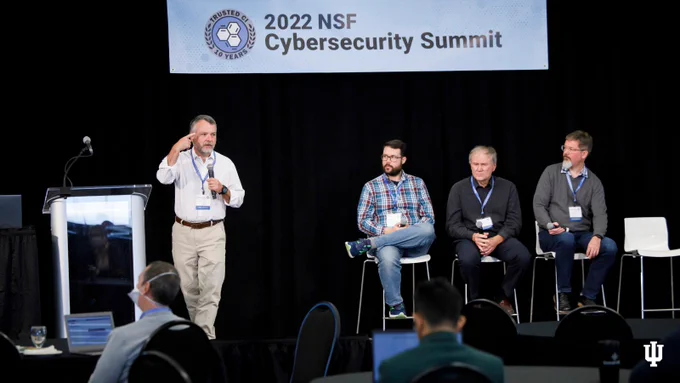 From the 2022 NSF Cybersecurity Summit at IU Bloomington. On the stage under a banner with the name of the event sit three figures on stools (right), looking towards the person giving a presentation (left). The presenter is holding a mic in one hand and gestures with the other, having stepped away from the glass podium (far left).