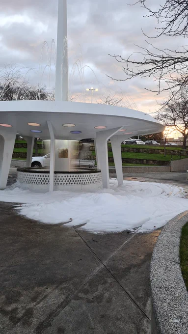 A white water fountain that looks a bit like a 1960s spaceship with lots of white foam from dish soap surrounding it on the ground.