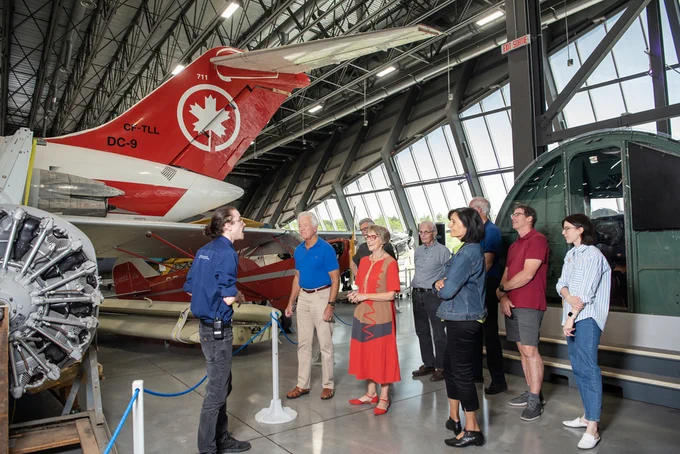A museum guide wearing a blue shirt and black pants delivers a tour to a dozen people who stand next to the tail of an Air Canada jet.