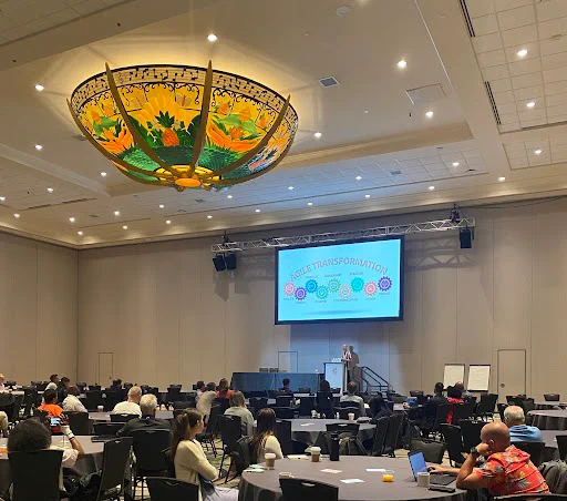 Photo of Dylan Barrell giving a talk with the slide agile transformation on screen in a ballroom