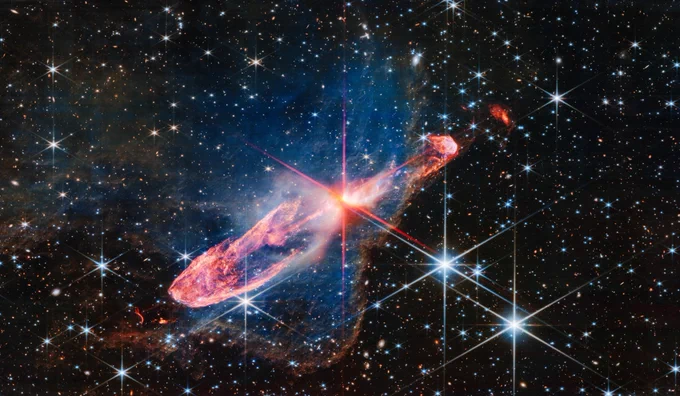 At the centre is a thin horizontal orange cloud tilted from bottom left to top right. It takes up about two-thirds of the length of this angle, but is thin at the opposite angle. At its centre is a set of very large red and pink diffraction spikes in Webb’s familiar eight-pointed pattern. It has a central yellow-white blob, which hides two tightly orbiting stars. The background is filled with stars and galaxies.