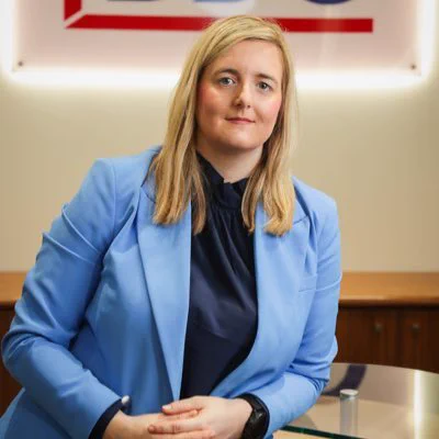 Lorraine Nelson LIB President headshot. Lorraine is wearing a navy top paired with a baby blue blazer. Hans crossed and leaning on a side table. Smiling. Background is beige.