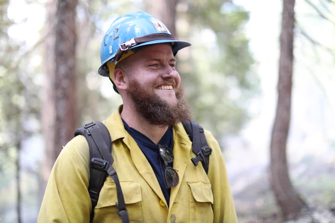 A firefighter wearing a hard hat and protective clothing stands and smiles while looking at the forest around him.